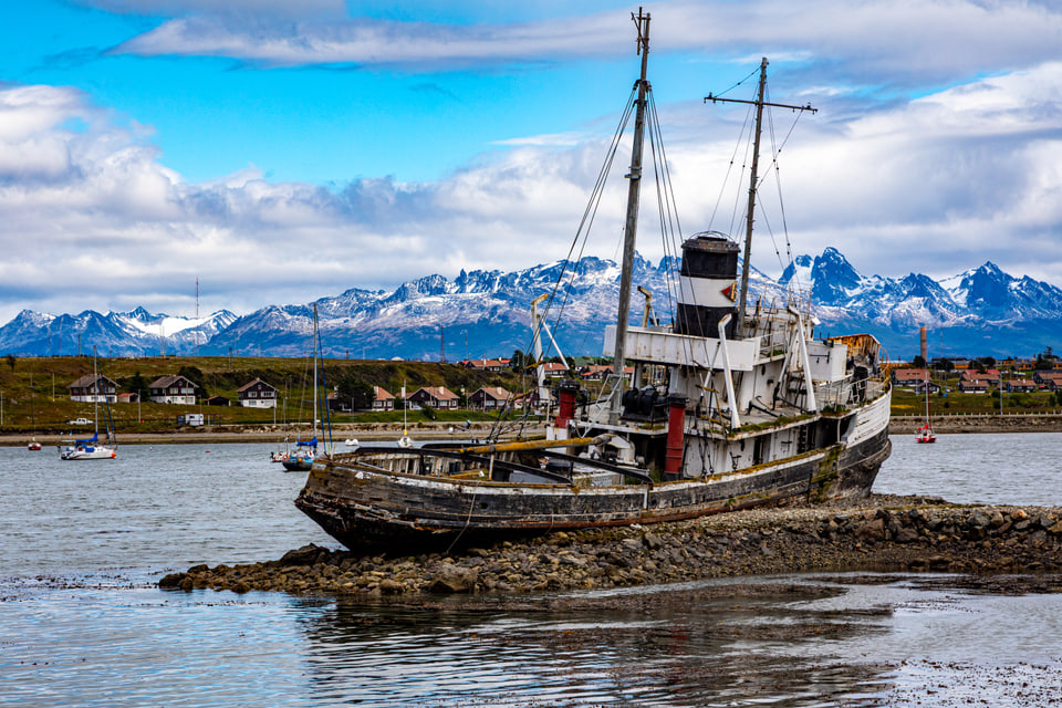 Weathered ship on a rocky shore with a town and snow-covered mountains beyond.