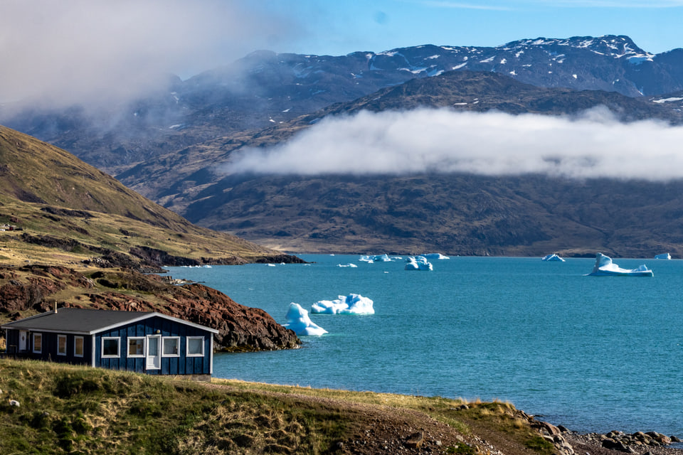 Blue house overlooking a fjord dotted with icebergs beneath misty mountains.